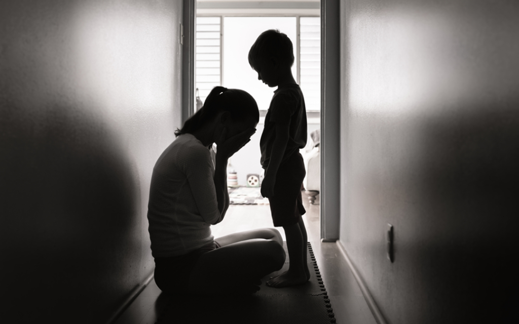 Struggling mother sits on floor holding head in hands, while young son stands next to her