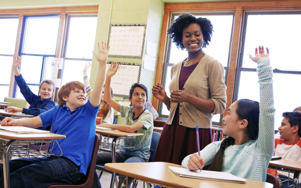 A black woman teacher smiles as she responds to an enthusiastic room of children.