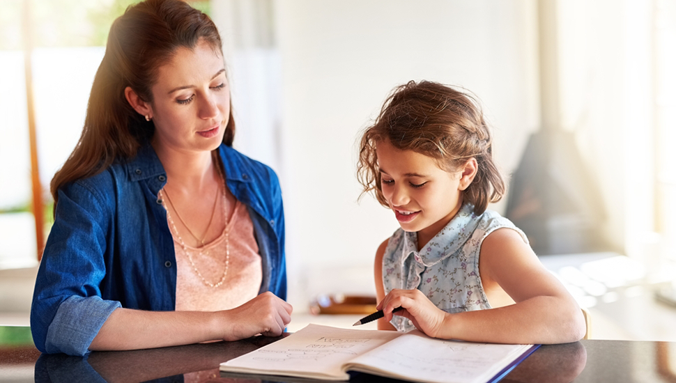 Mom and preteen daughter sit together at a table reading from notebook