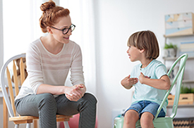 Smiling little boy talking with cheerful child psychotherapist during therapy session at office