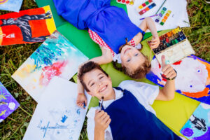 Smiling boy and girl laying down, surrounded by art supplies and paintings