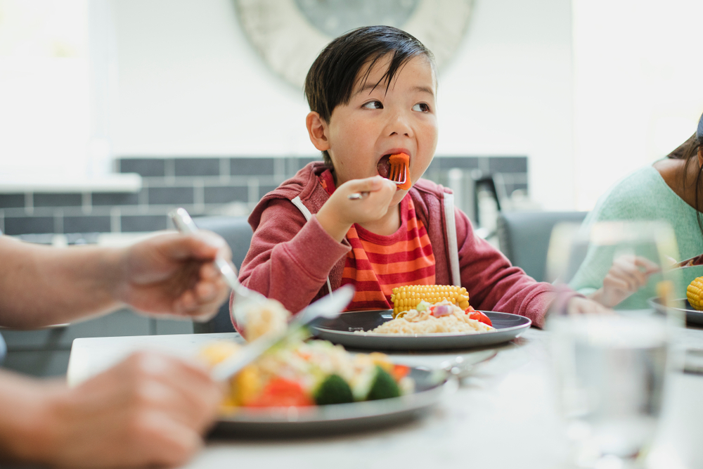 Little boy eating dinner with family, having a mouthful of veggies.