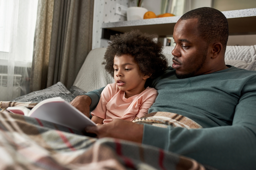 Focused,Adult,Father,And,Little,Son,Reading,Book,On,Sofa